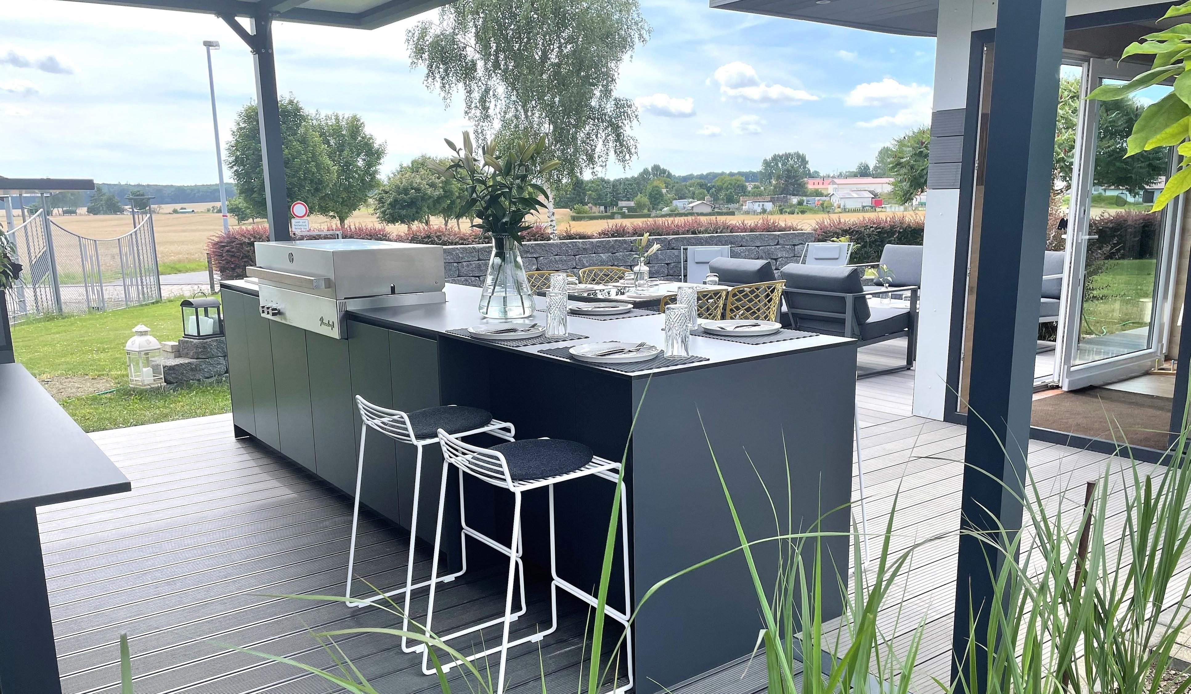Moderne Outdoor-Küche mit Barhockern auf einer Terrasse, Ausblick auf Felder und Wiese.