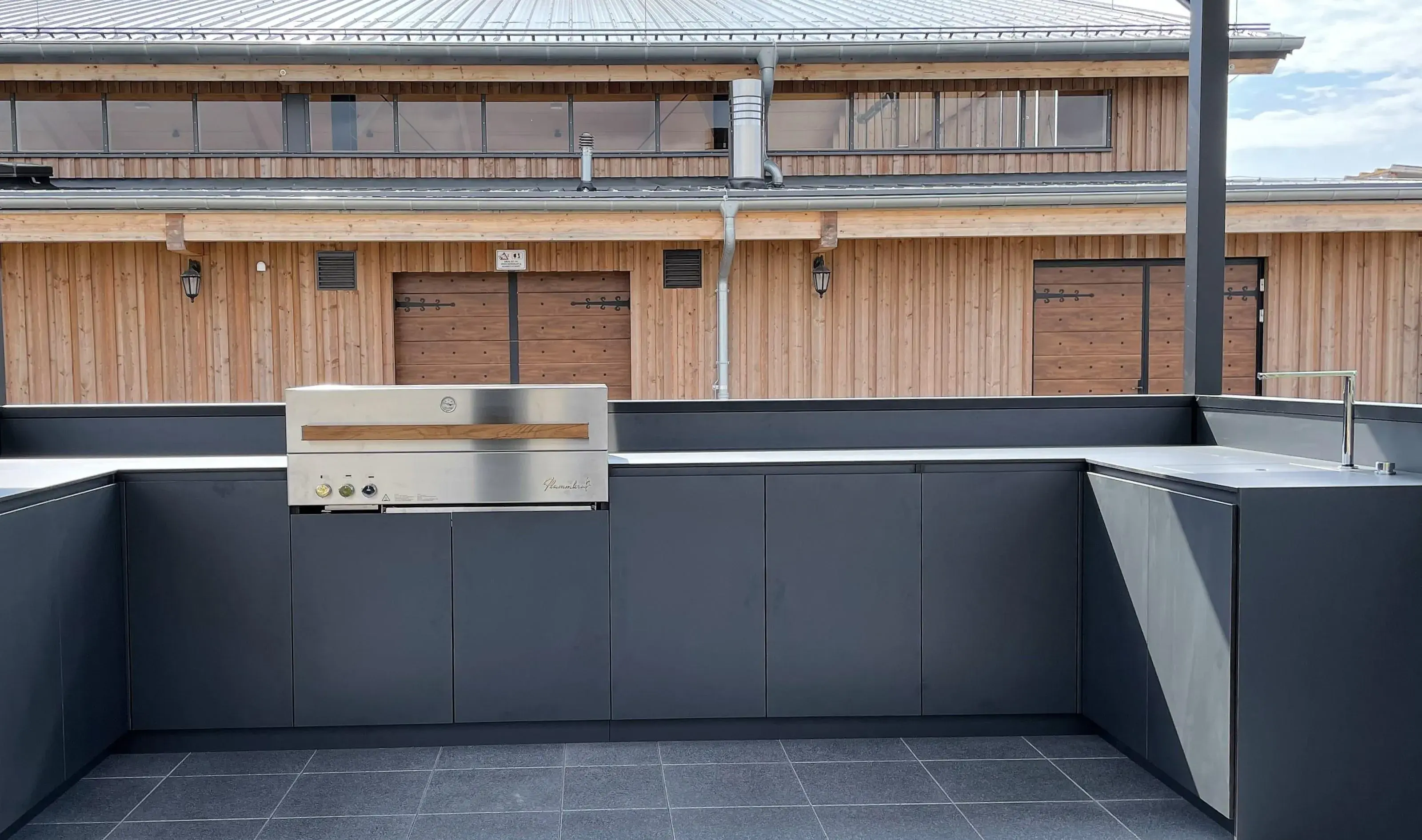 Moderne Außenküche mit dunklen Schränken und integriertem Grill vor Holzgebäude