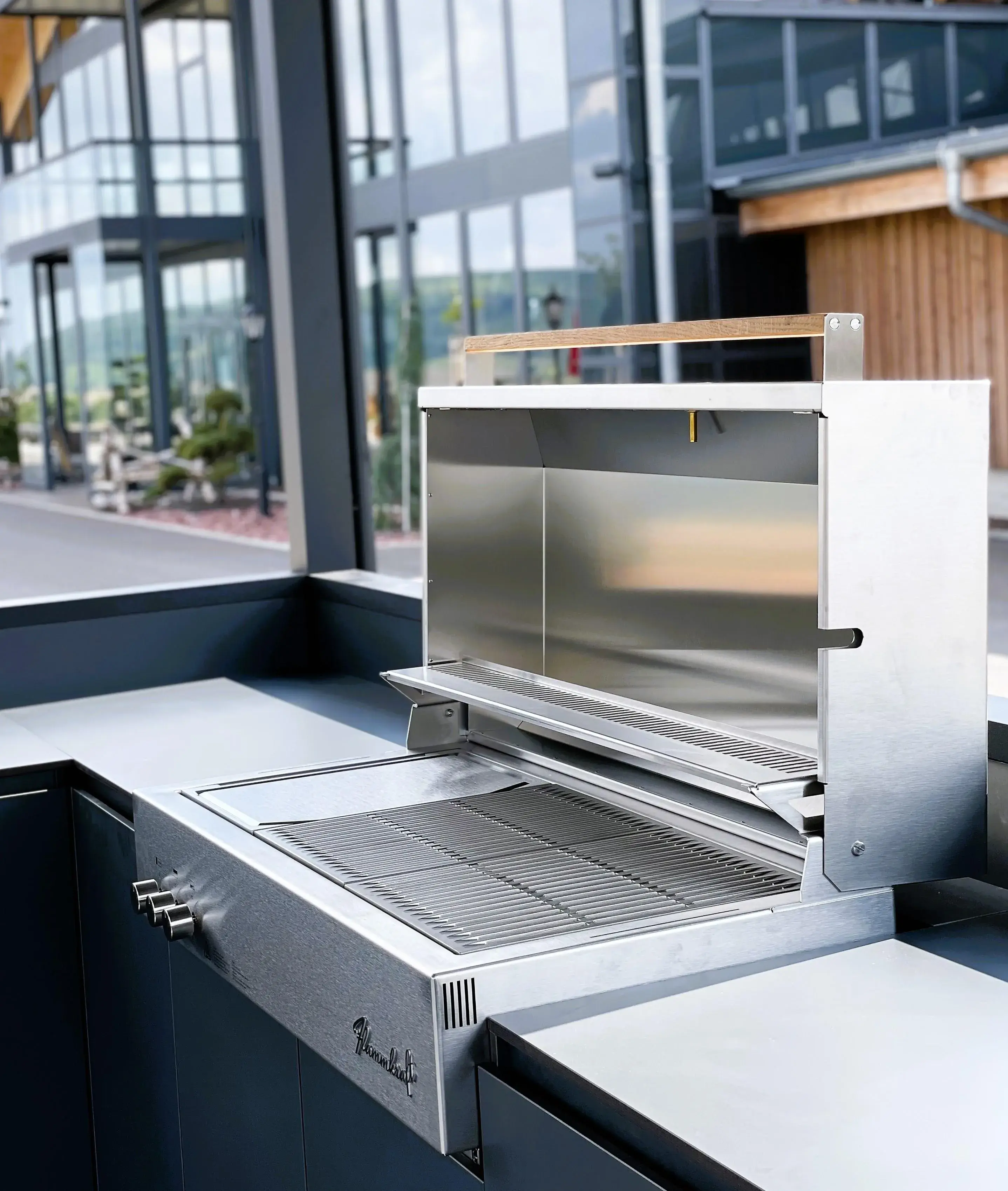 Silberner Grill mit offenem Deckel auf moderner Terrasse, Gebäude im Hintergrund.