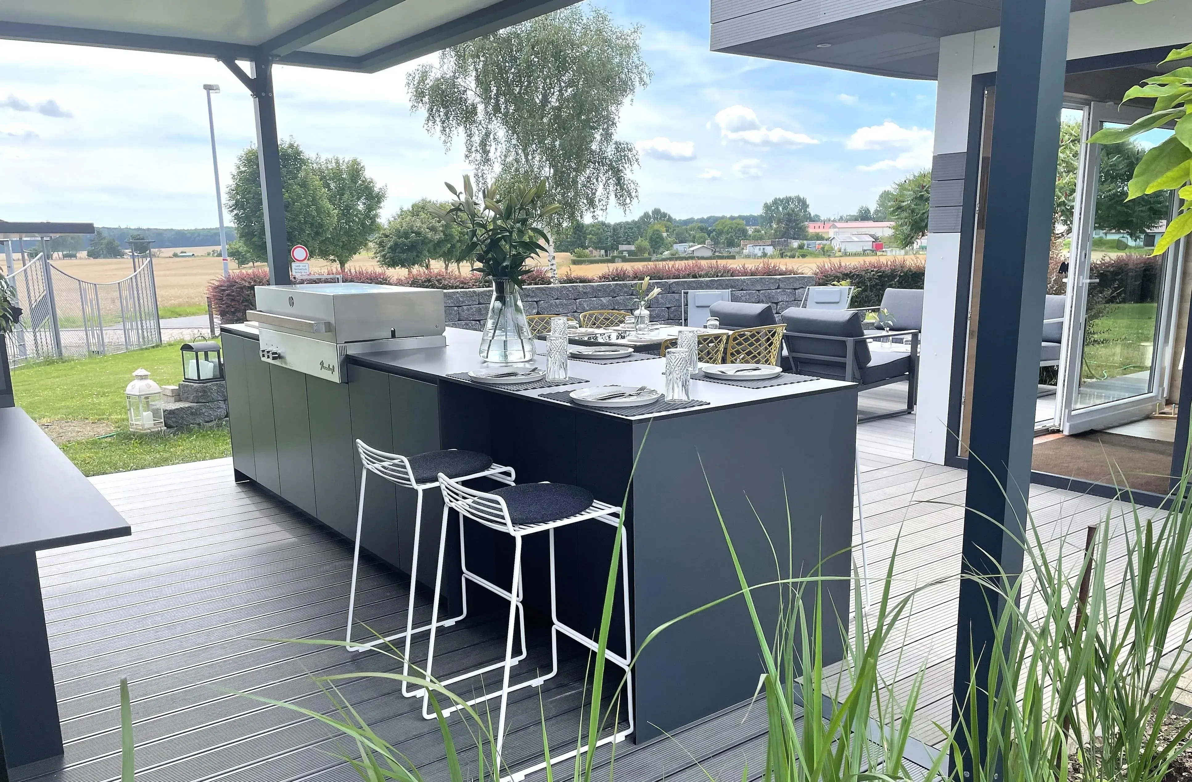 Moderne Outdoor-Küche mit Barhockern auf einer Terrasse, Ausblick auf Felder und Wiese.