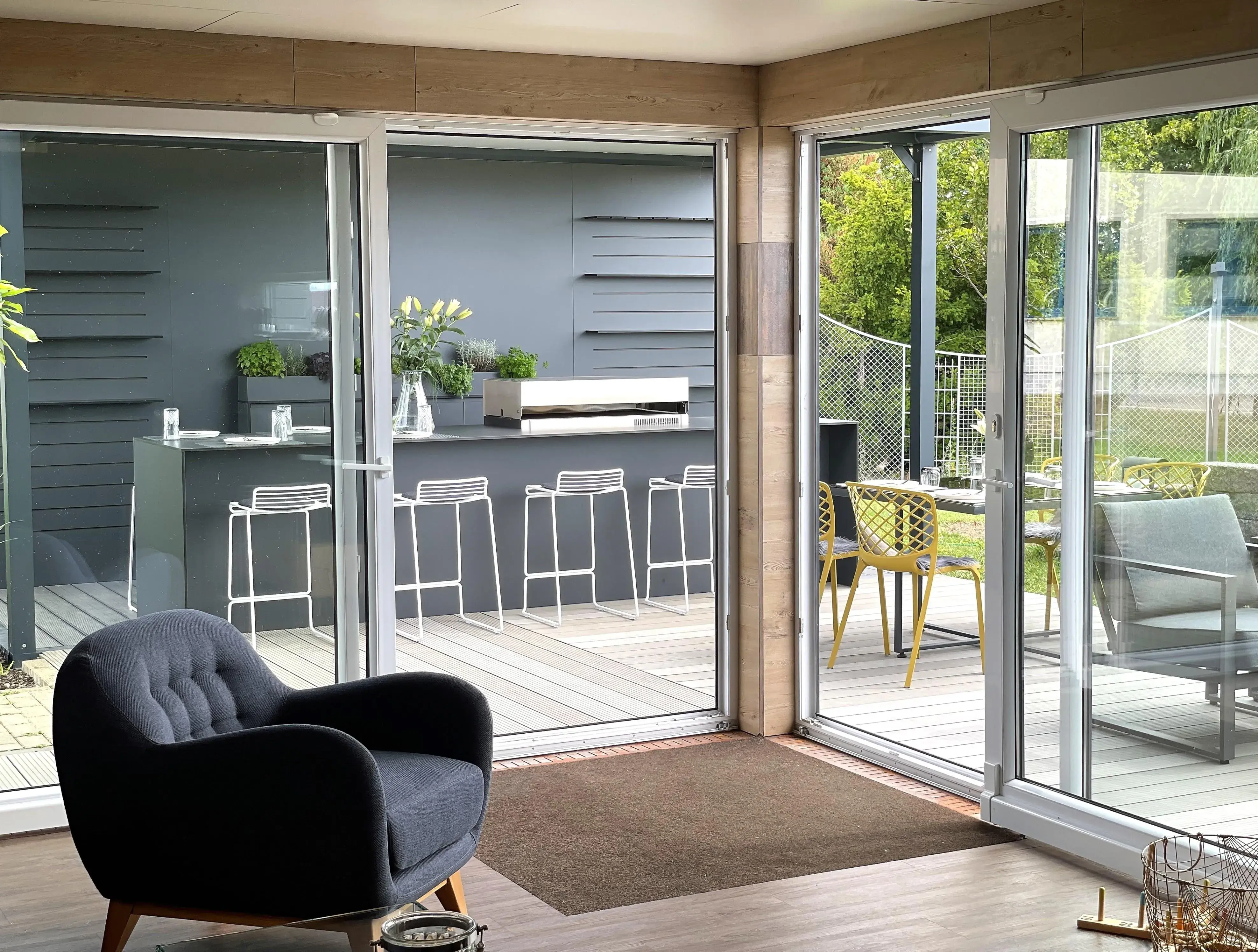 Heller Raum mit Sessel, Glaswänden und Terrasse mit Barhockern und Pflanzen.
