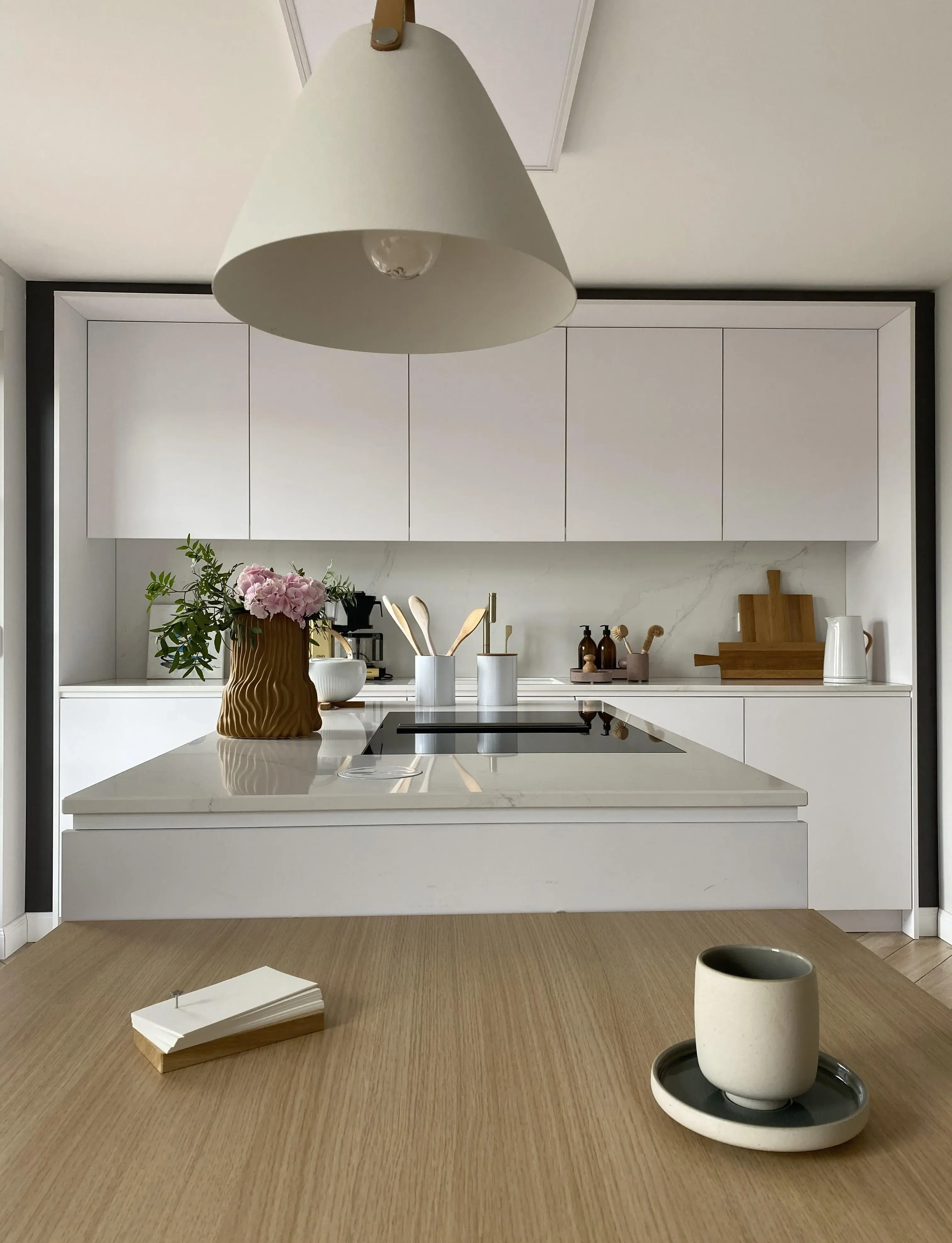 Moderne weiße Küche mit Kochinsel, Vase mit Blumen, Tasse und Notizblock auf Tisch.