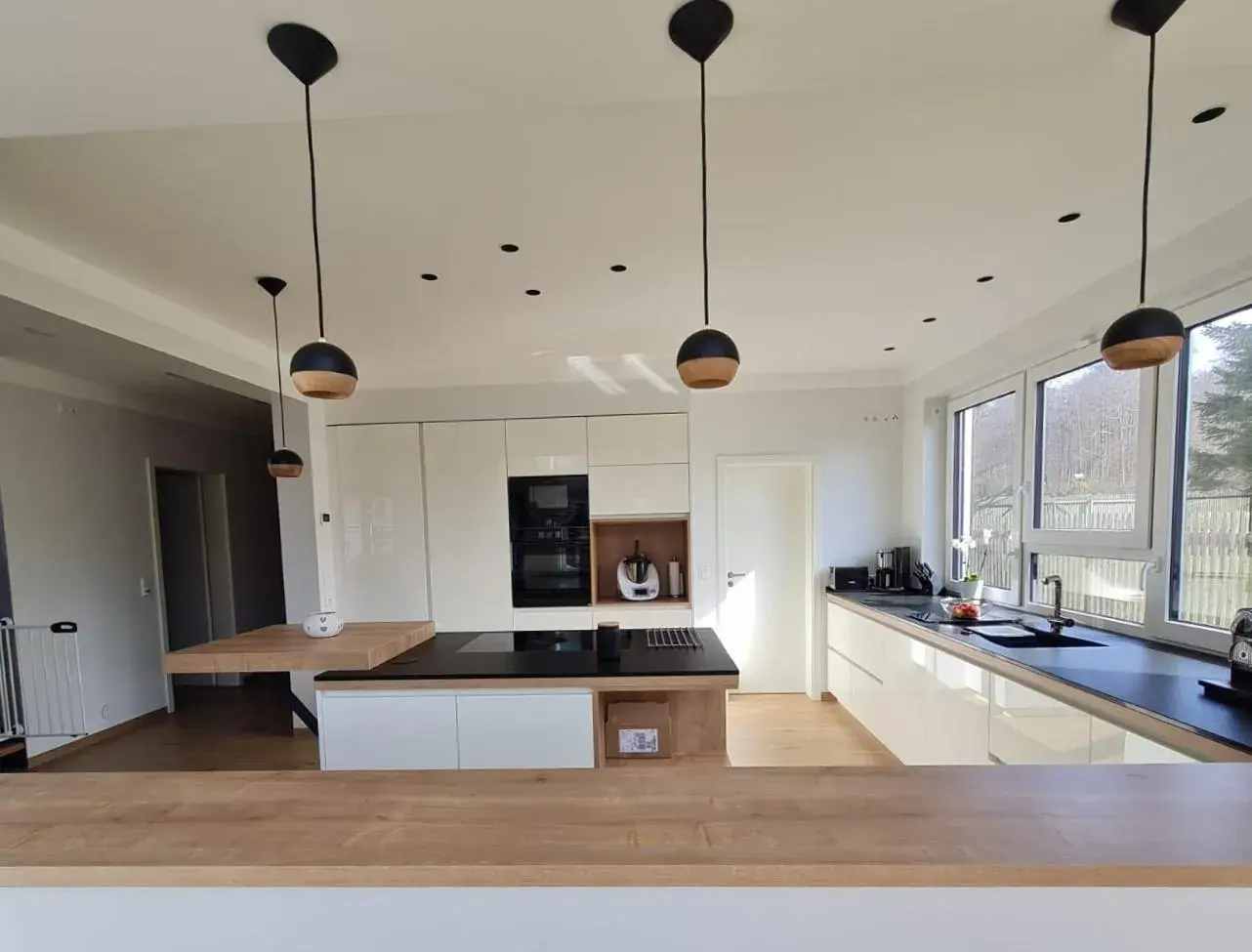 Moderne, helle Küche mit Holz- und weißen Oberflächen, großen Fenstern und Hängelampen.