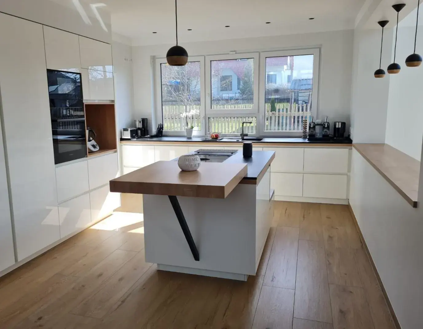 Moderne weiße Küche mit Holzarbeitsplatten, Insel und großen Fenstern, viel Tageslicht.